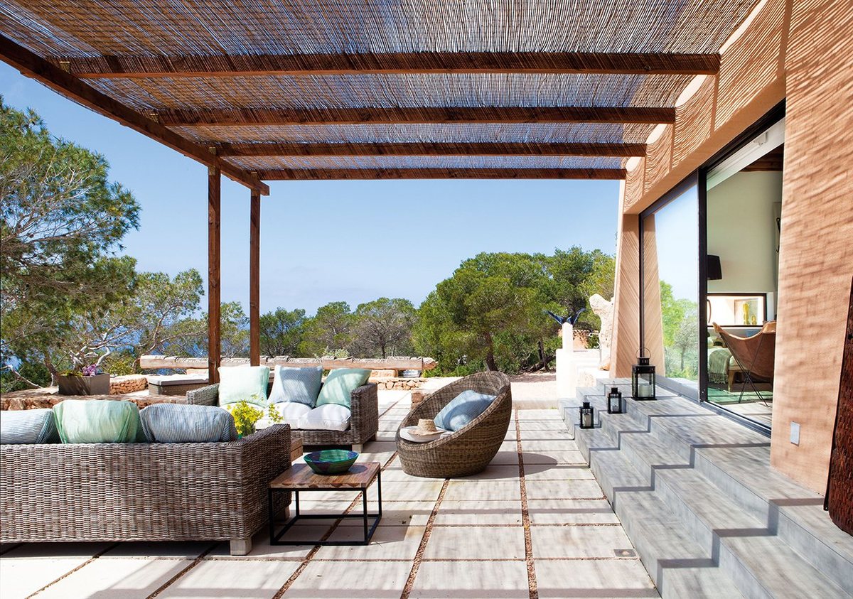 Своеобразная летняя гостиная на террасе под тростниковой крышей с плетеной мебелью.