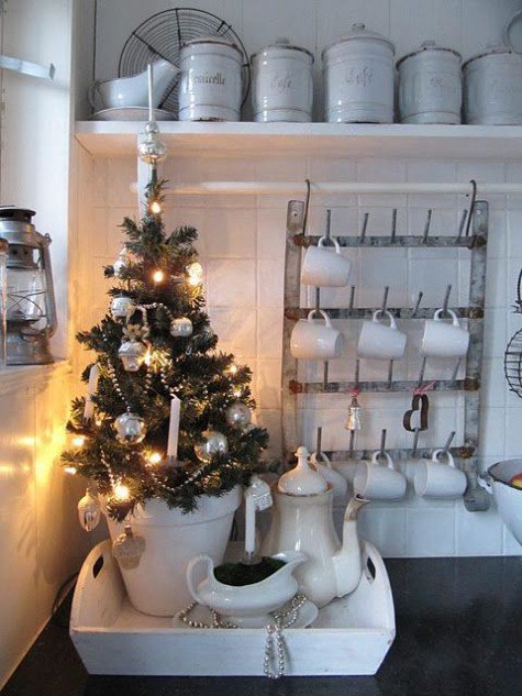 Крошечная елка в горшке на подносе украшает кухню. На елке серебристые шарики и светящаяся гирлянда.